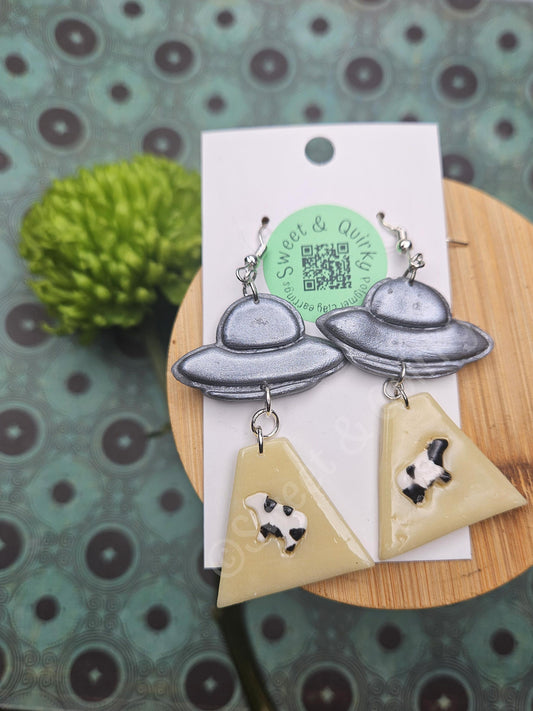 Earrings with UFO and a cow being abducted designs on a card with a green plant in the background