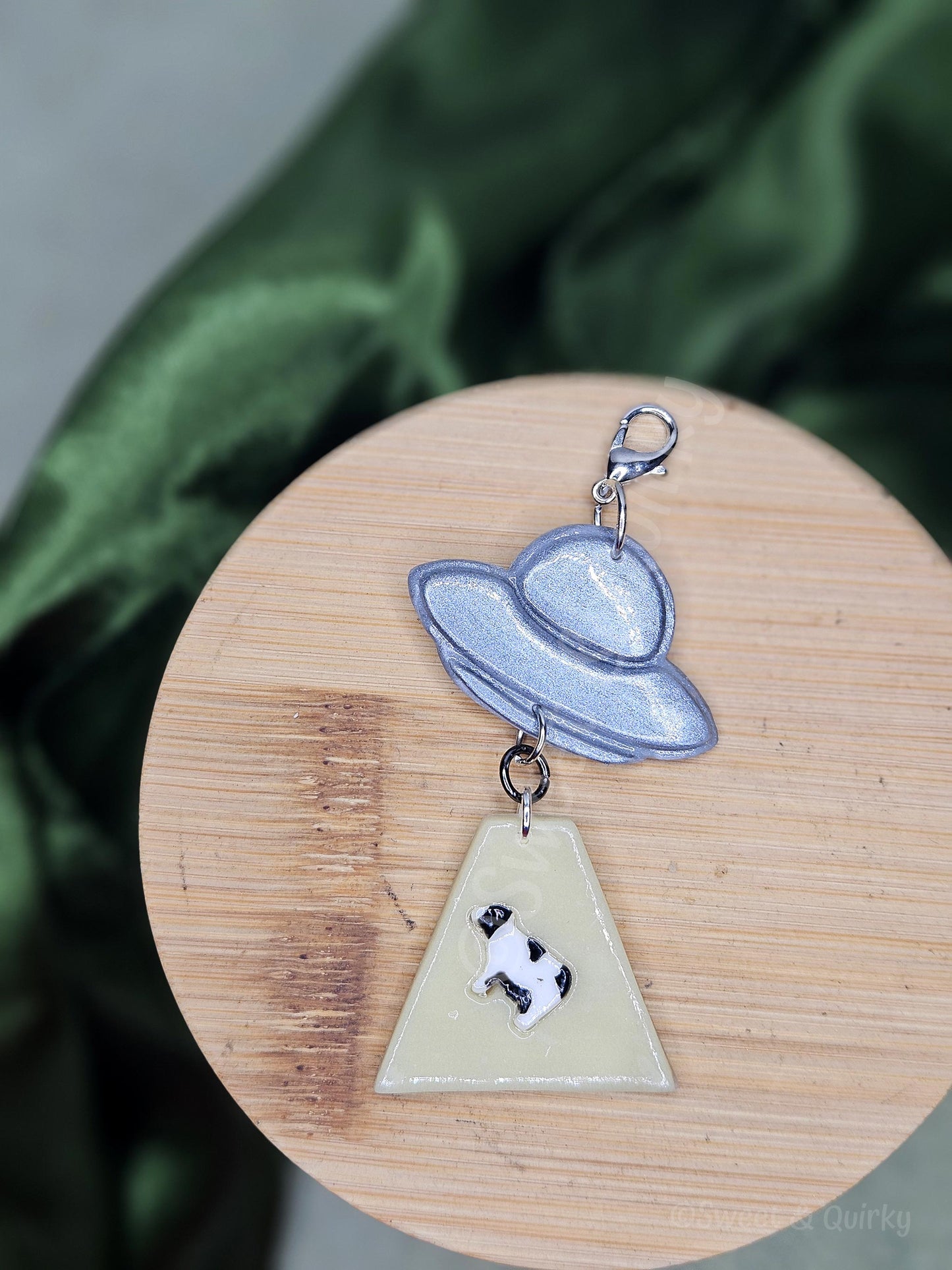 Lobster clasp charm with an abducted cow and spaceship design on a wooden surface