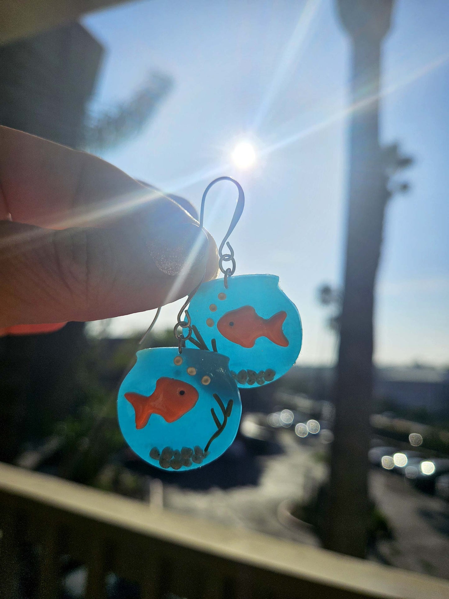 Earrings with goldfish design held against a blurred outdoor background