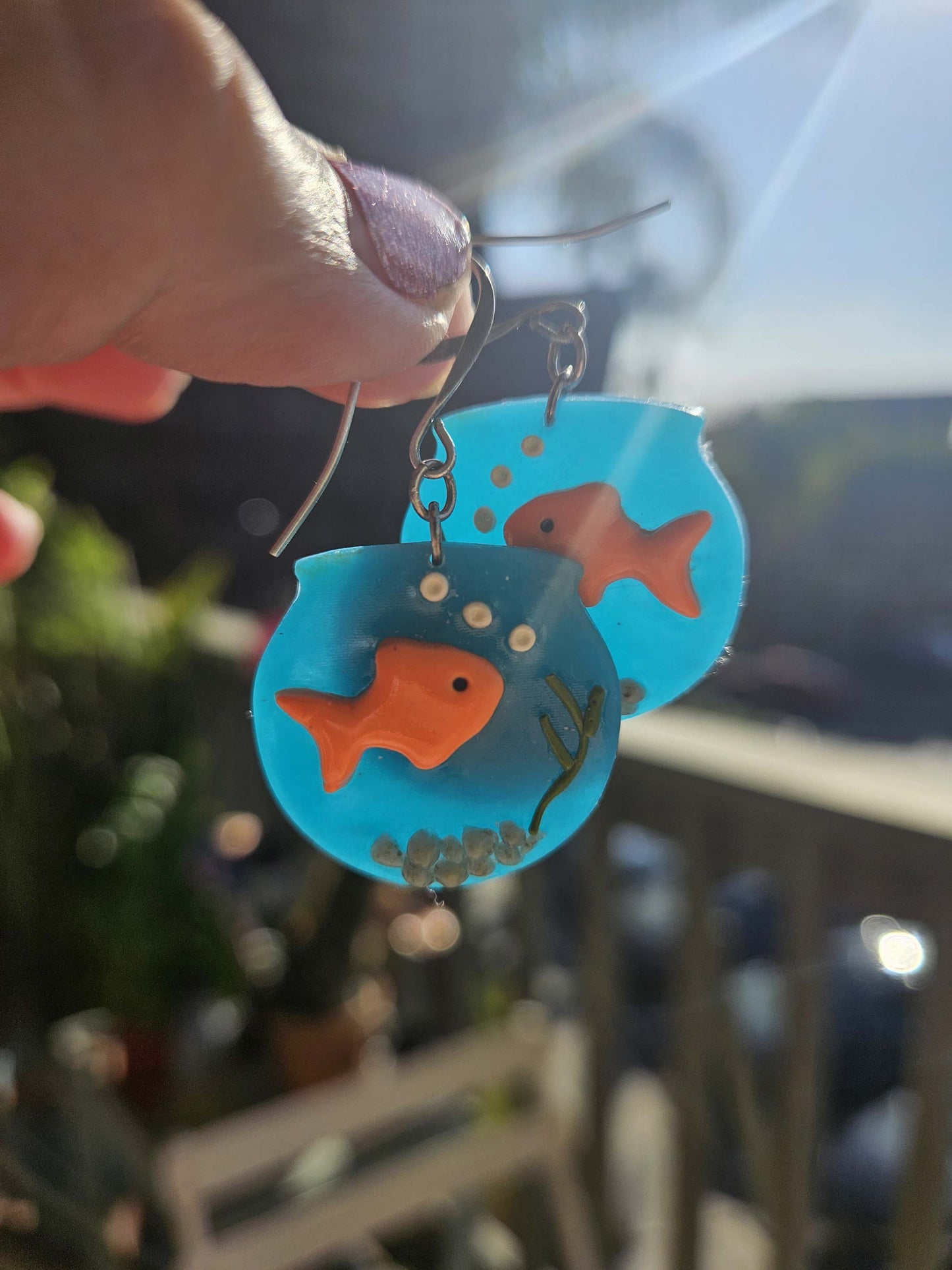 Earrings with goldfish design held by a hand against a blurred outdoor background