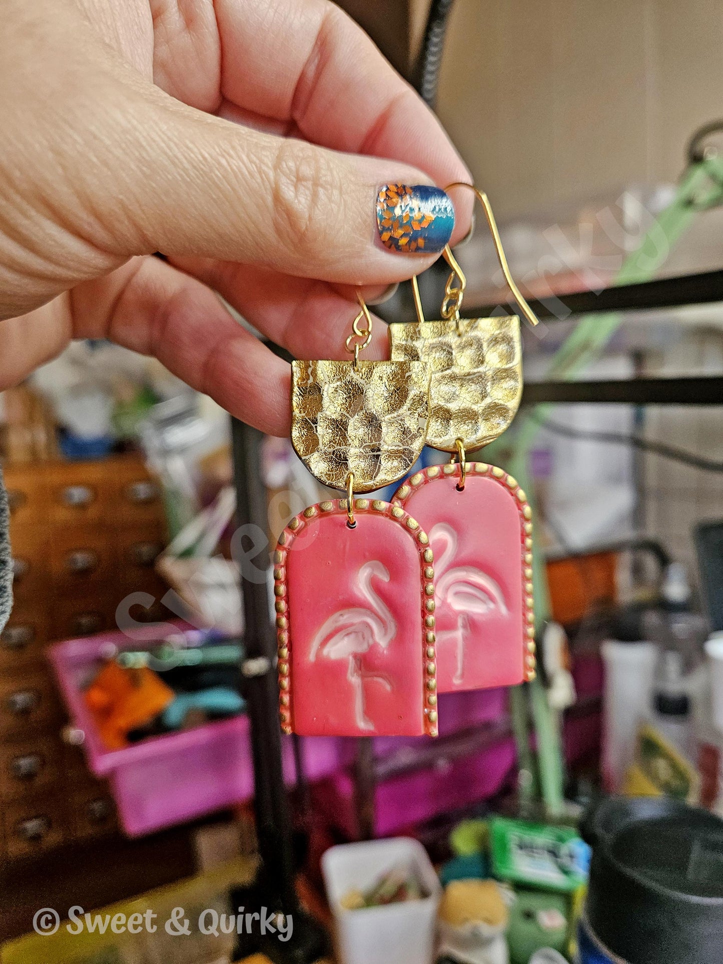 Gold and pink earrings with flamingo design held by a hand in a cluttered room.
