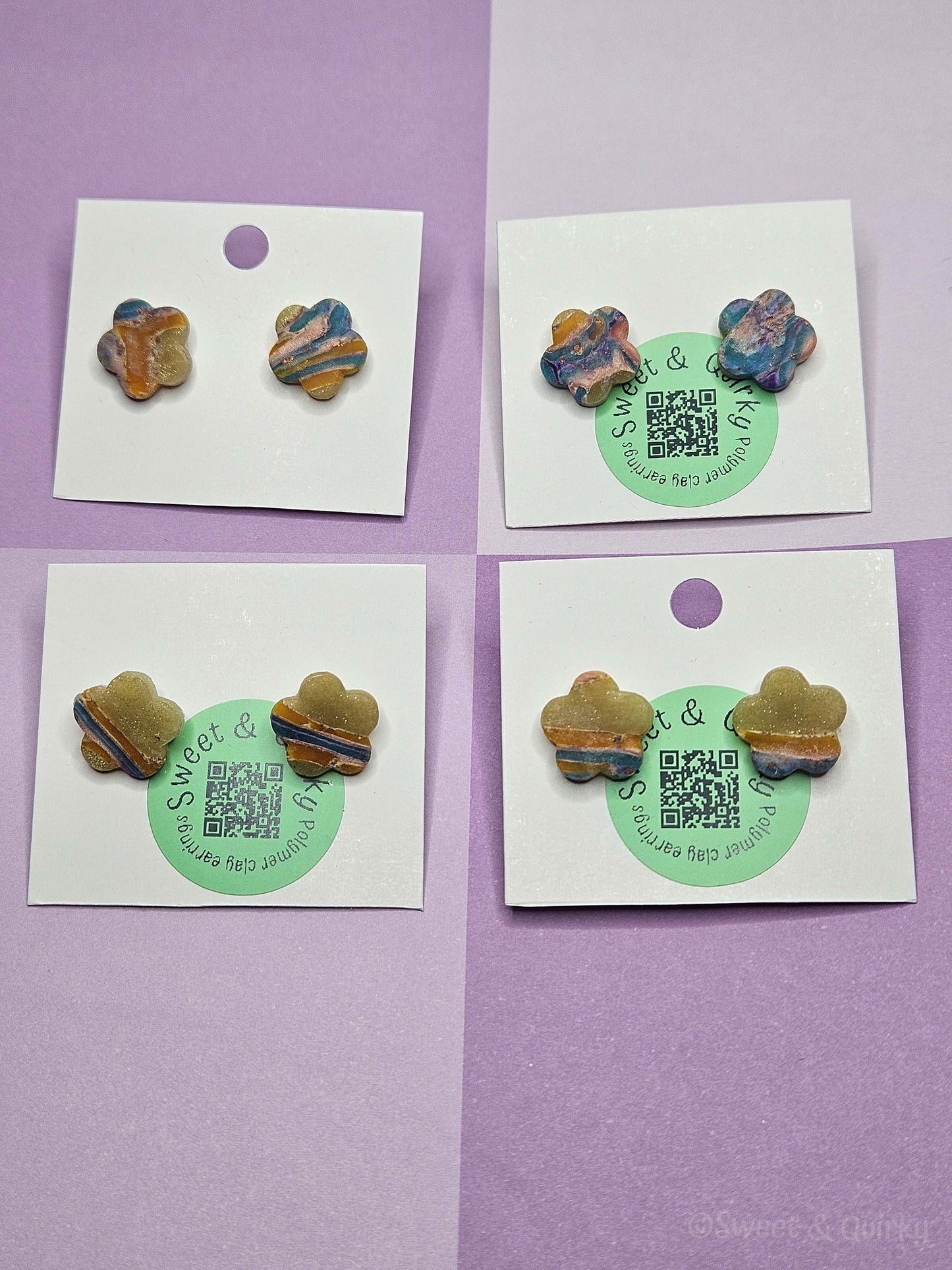 Four pairs of colorful clay earrings on white cards with green labels against a purple background.