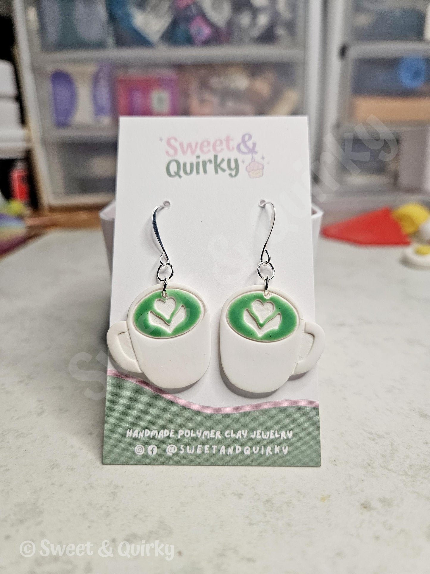 Earrings shaped like coffee cups with green designs on a Sweet & Quirky branded card.