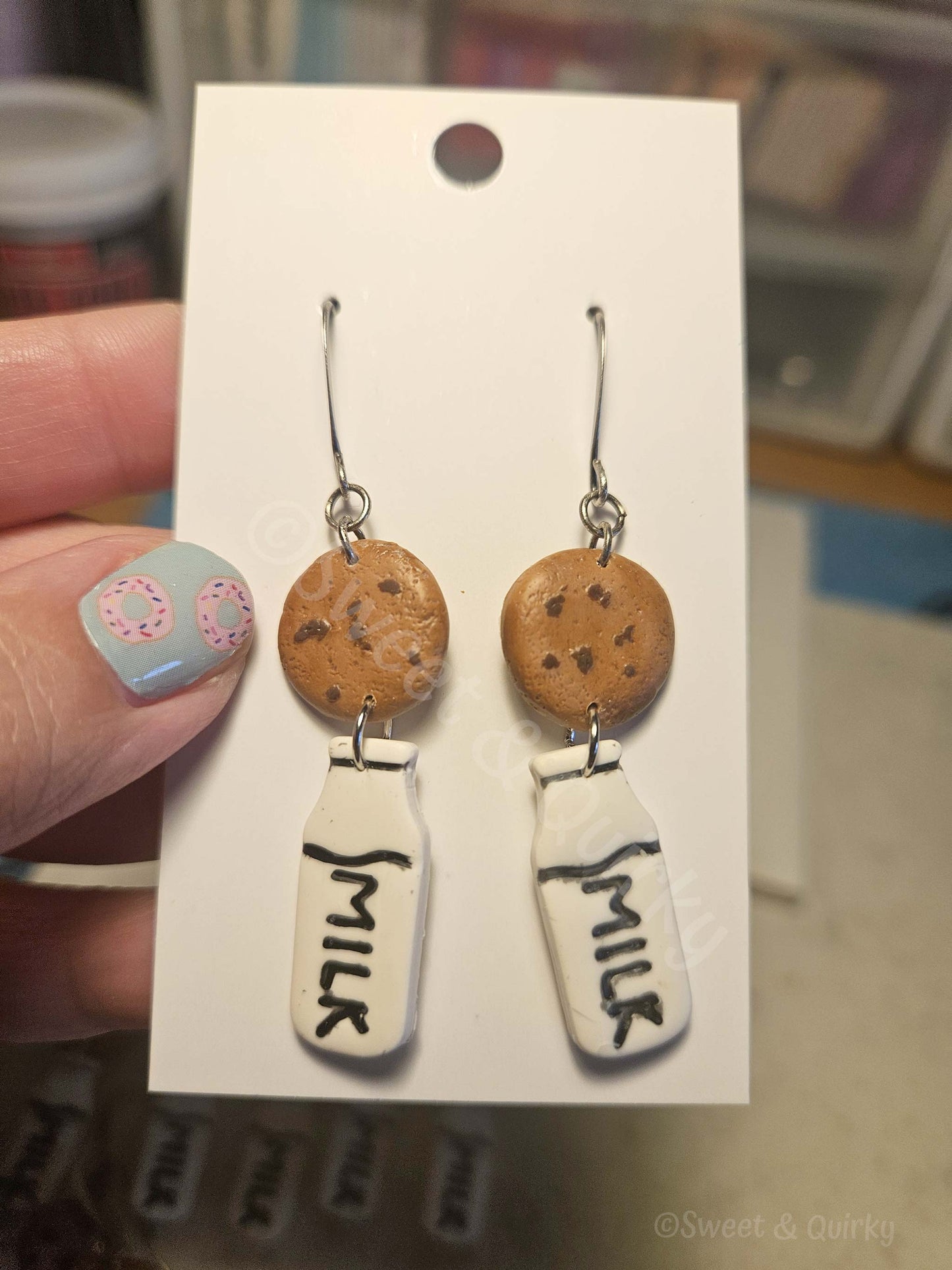 Cookie and milk earrings on a display card held by a hand with nail polish.