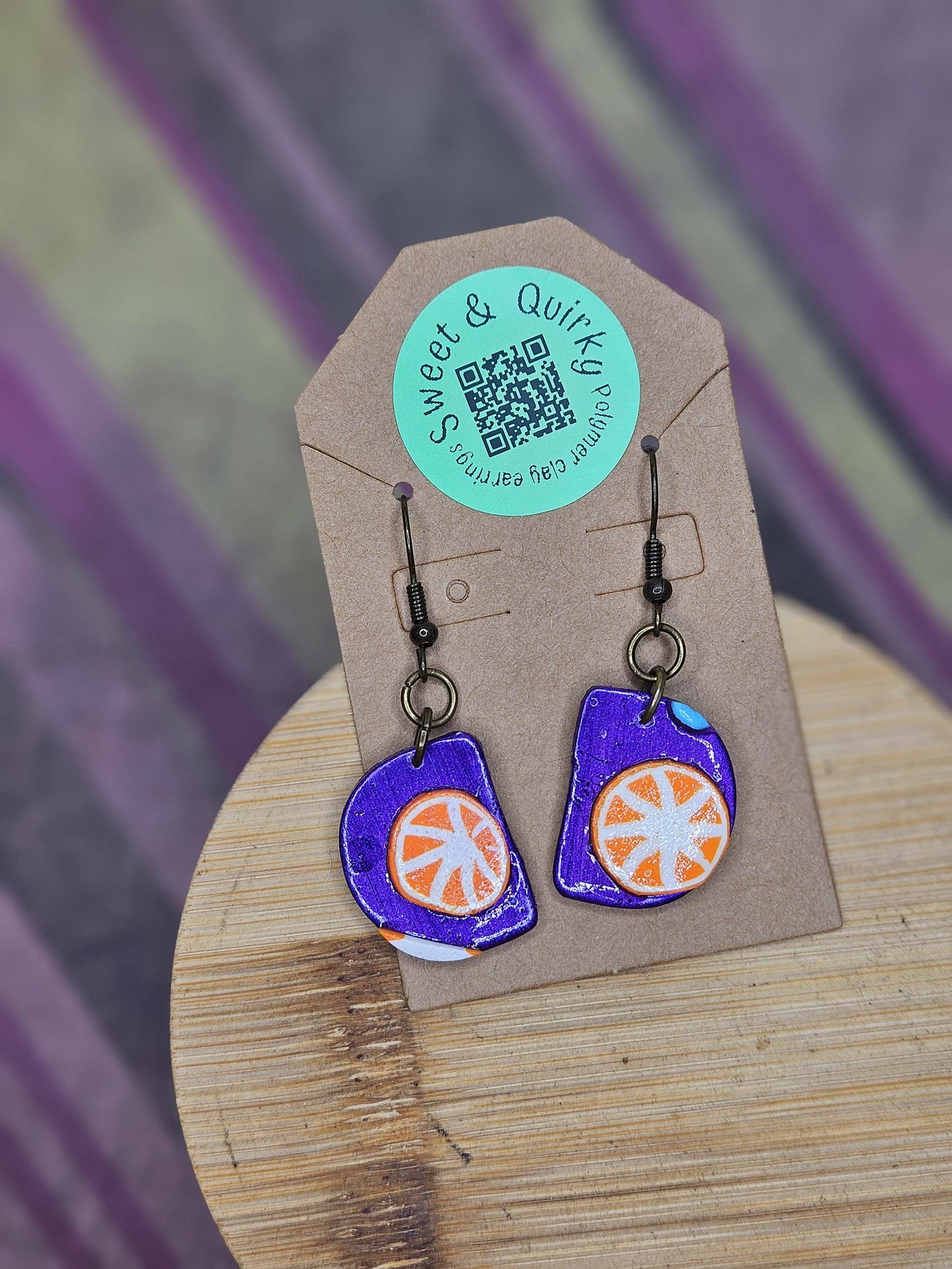 Pair of purple and orange earrings on a Sweet & Quick branded card with a blurred background
