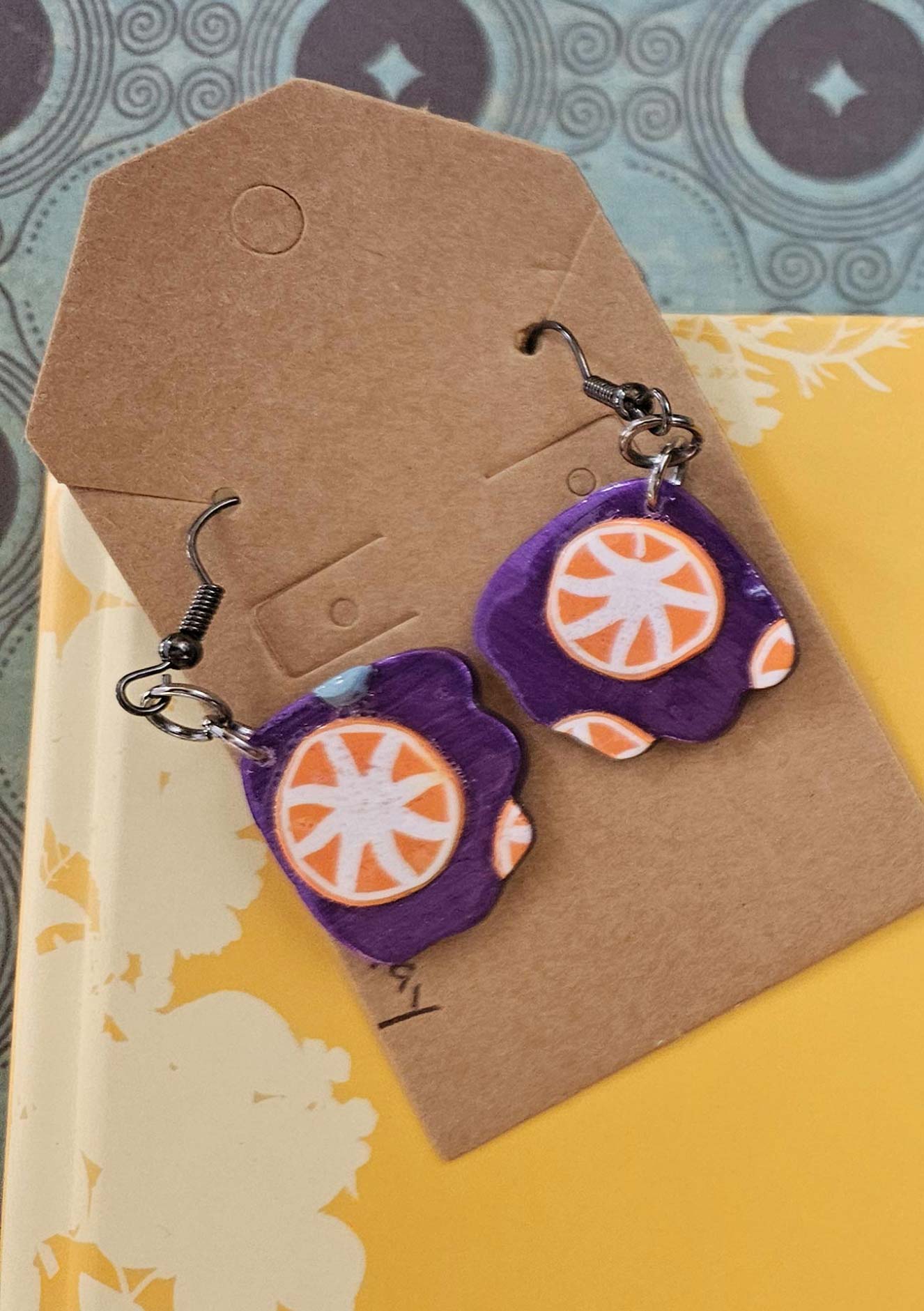 Pair of purple earrings with orange circular designs on a brown card.