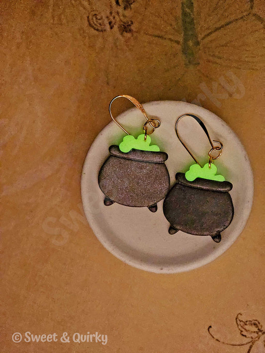 Pair of cauldron-shaped earrings with green accents on a beige background.