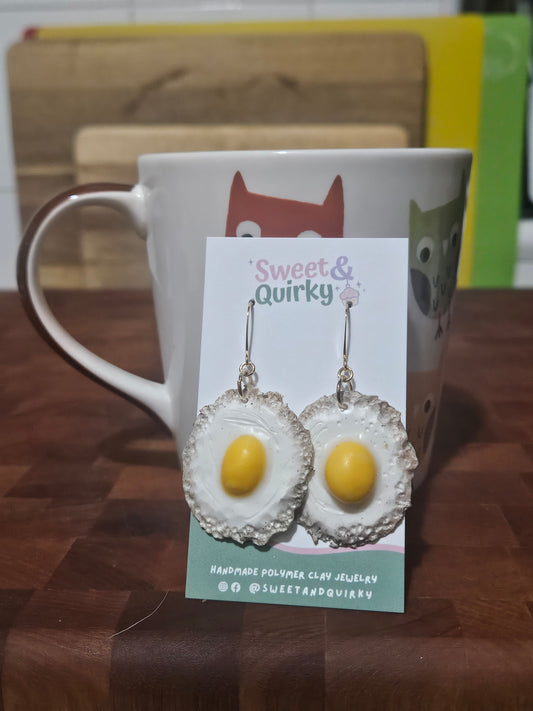 Earrings shaped like fried eggs on a 'Sweet & Quirky' branded card, with a mug in the background.