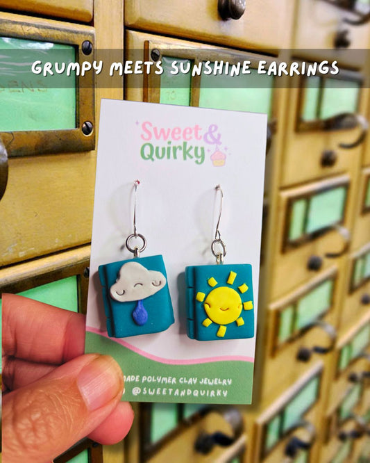 A pair of handmade clay earrings displayed on a card held by a hand. The left earring is a teal square with a white cloud charm dripping a blue tear, and the right earring is a teal square with a yellow smiling sun.