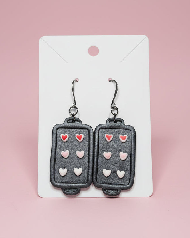 Earrings shaped like small baking sheets with heart sugar cookies on a white card.