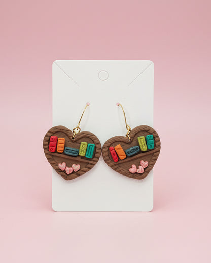 Wooden bookshelf earrings with books and hearts on a card with a light pink background.