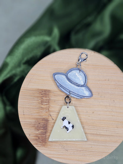 Lobster clasp charm with an abducted cow and spaceship design on a wooden surface