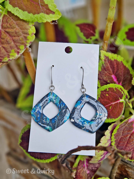 Blue and silver square earrings on a white card with green and red leaves in the background