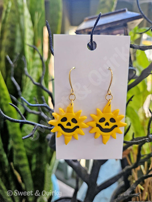 Yellow sun-shaped earrings with black smiley face on a white card against a blurred green plant background.