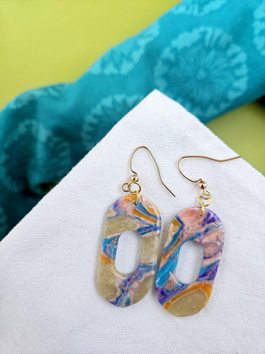 Colorful marbled earrings on a white triangle with a blue fabric background