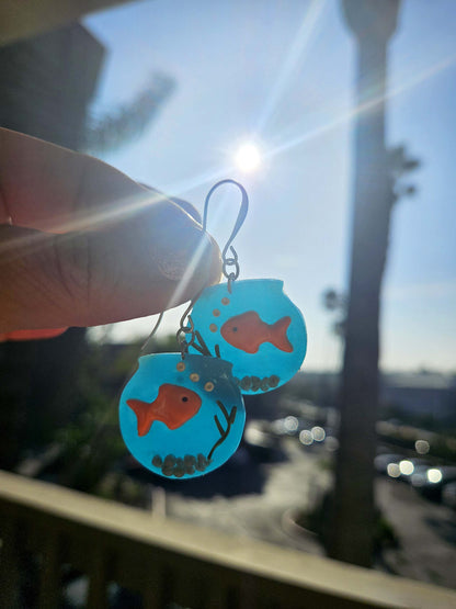 Earrings with goldfish design held against a blurred outdoor background