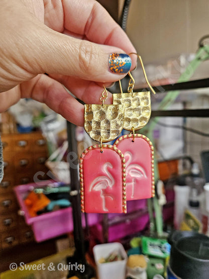 Gold and pink earrings with flamingo design held by a hand in a cluttered room.