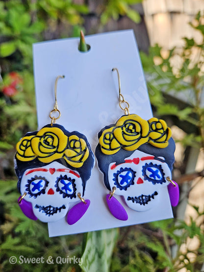 Colorful sugar skull earrings with yellow roses on a card with a green leafy background.