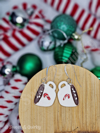 Pair of earrings shaped like white mugs with brown handles on a wooden stand, with Christmas ornaments in the background.