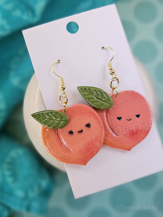 Peach-shaped earrings with facial expressions on a white card against a teal background
