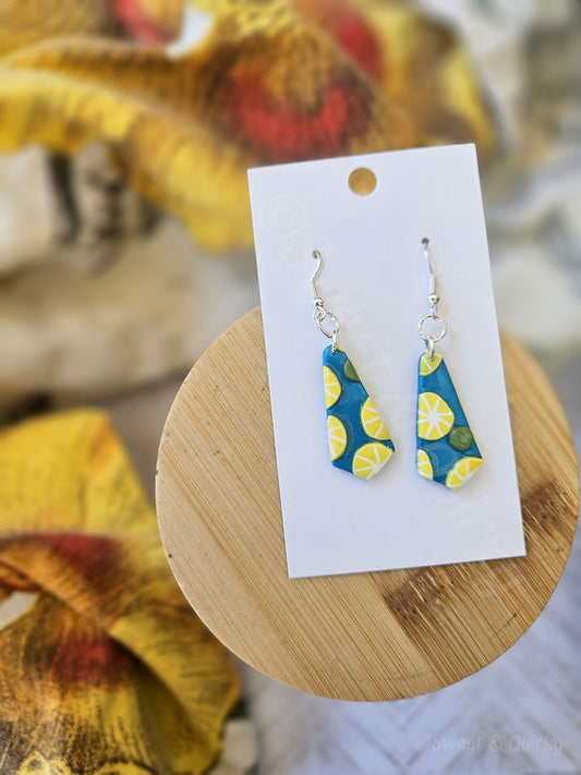 Pair of blue and yellow earrings on a white card with a wooden stand, floral background. Earrings feature lemon slices and green dots.