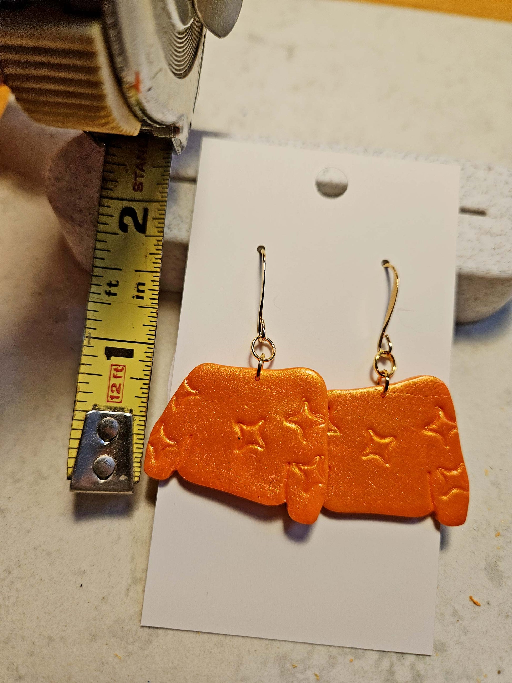 Orange earrings with star patterns on a white card, next to a ruler for scale.