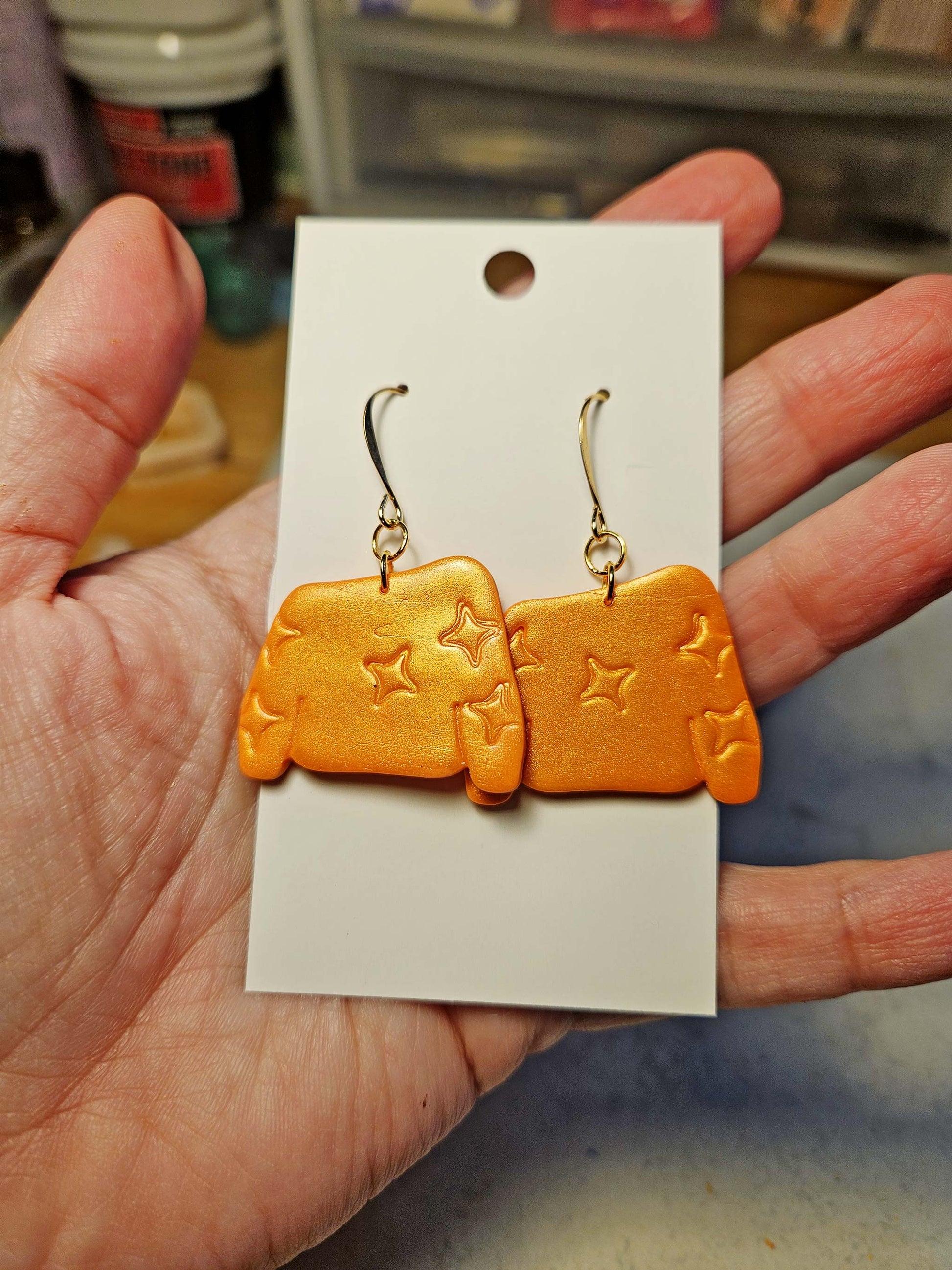 Orange sweater earrings with star designs on a white card held by a hand.