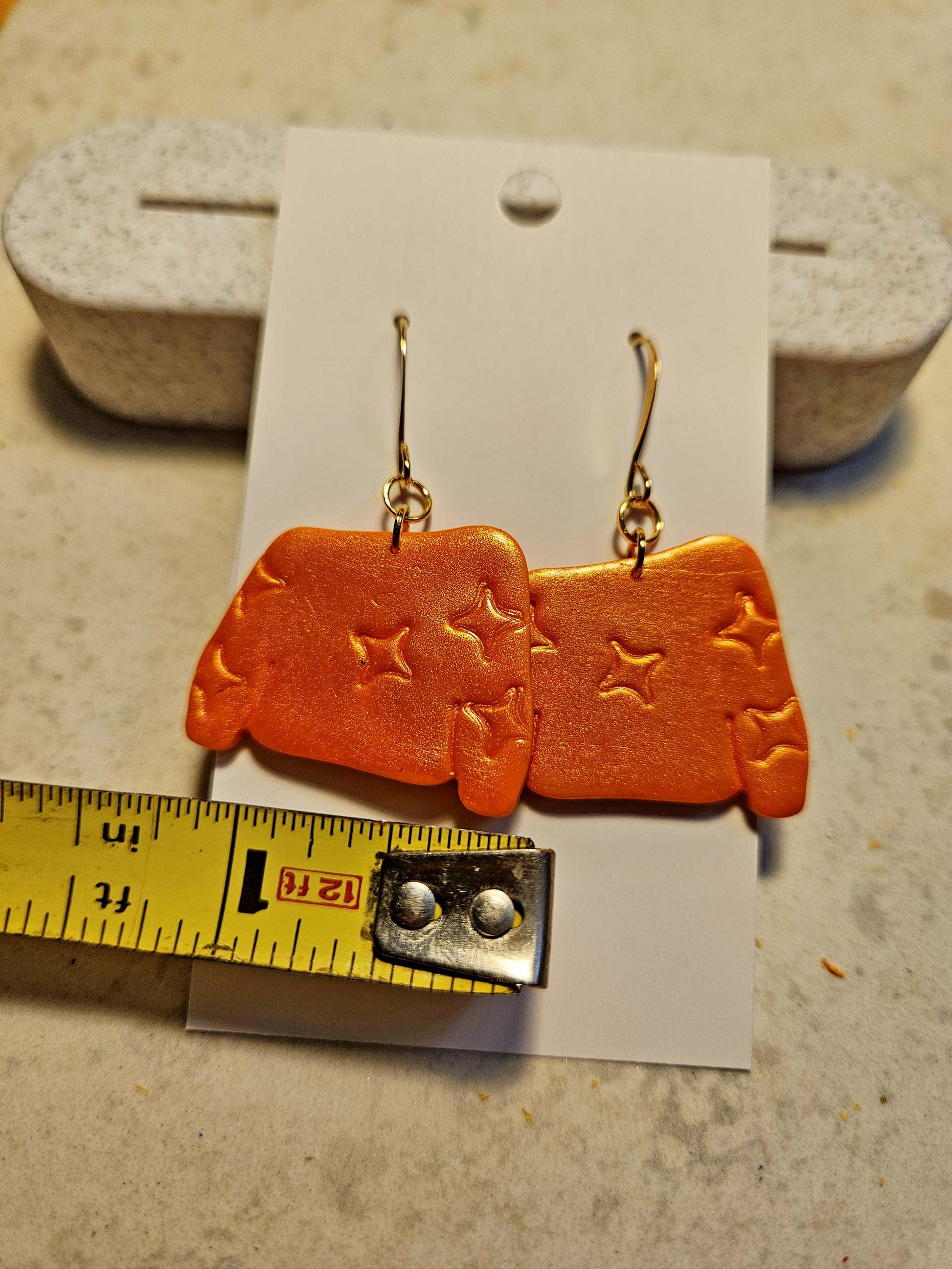 Orange earrings with star designs on a card, with a ruler for scale.