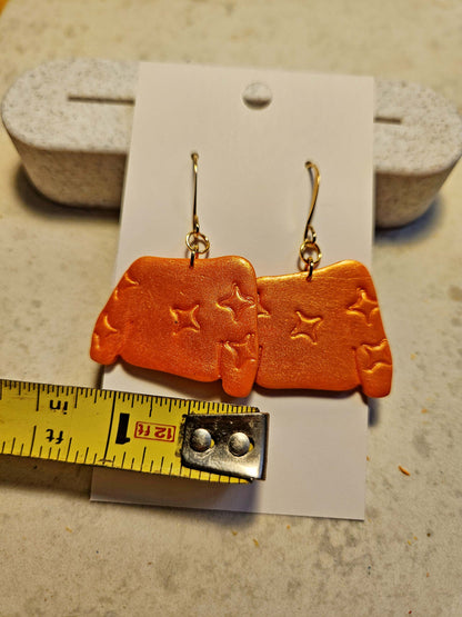 Orange earrings with star designs on a card, with a ruler for scale.