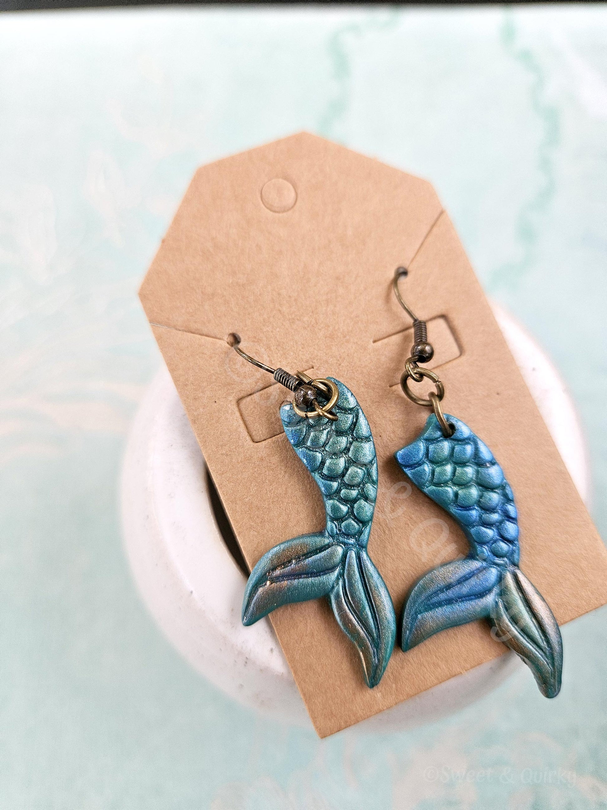Blue and green mermaid tail earrings on a cardboard backing with a light blue background