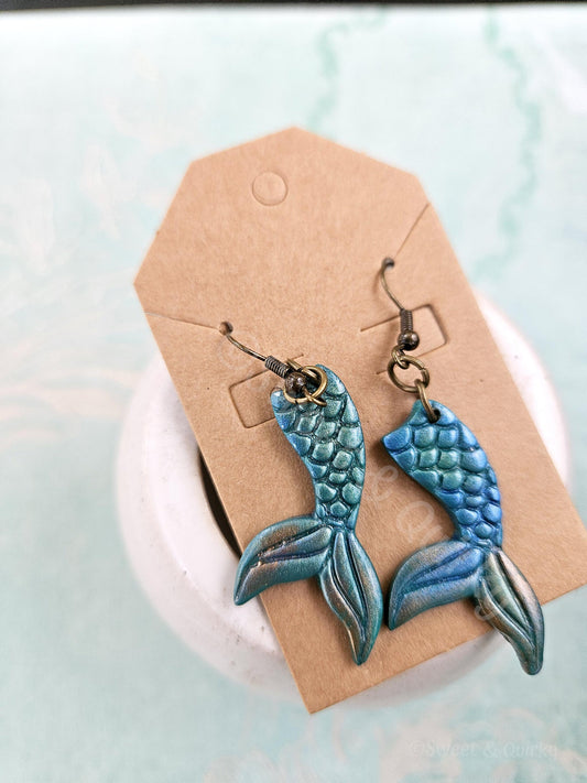 Blue and green mermaid tail earrings on a cardboard backing with a light blue background