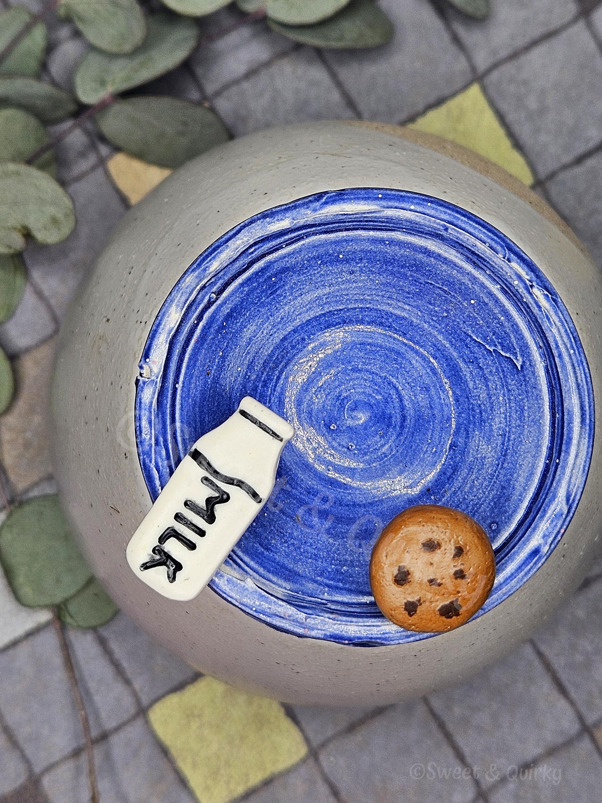 Ceramic jar with blue interior, milk carton label, and cookie on a textured surface. Stud earrings.