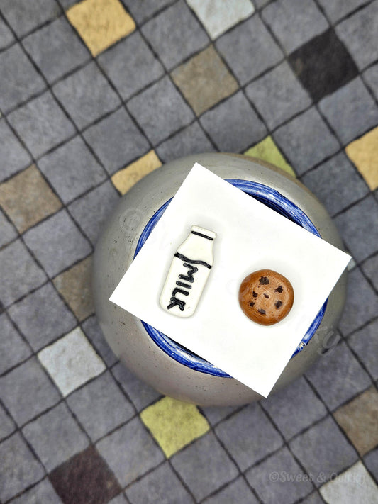 Small cookie and milk bottle stud earings on a white card with a checkered background.