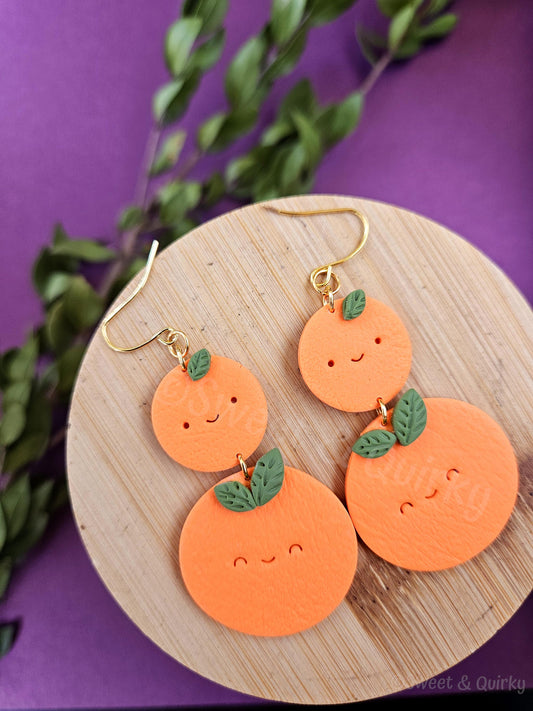 Orange fruit-shaped earrings with smiley faces on a wooden board against a purple background.