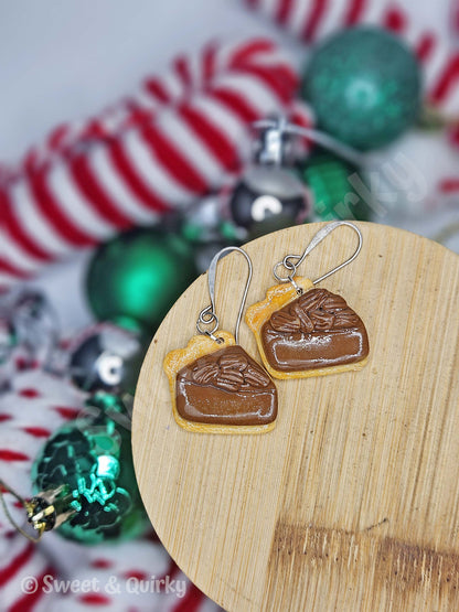 Earrings shaped like pecan pie on a wooden board, with Christmas decorations in the background.