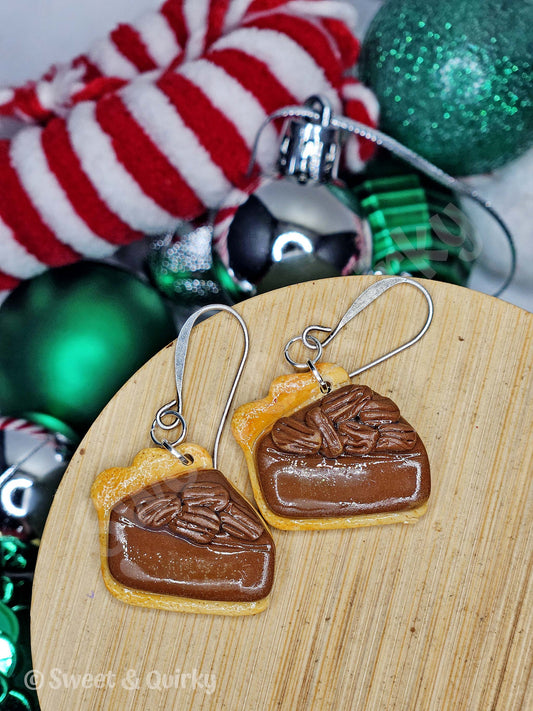 Pecan pie-shaped earrings on a wooden board with Christmas ornaments in the background.