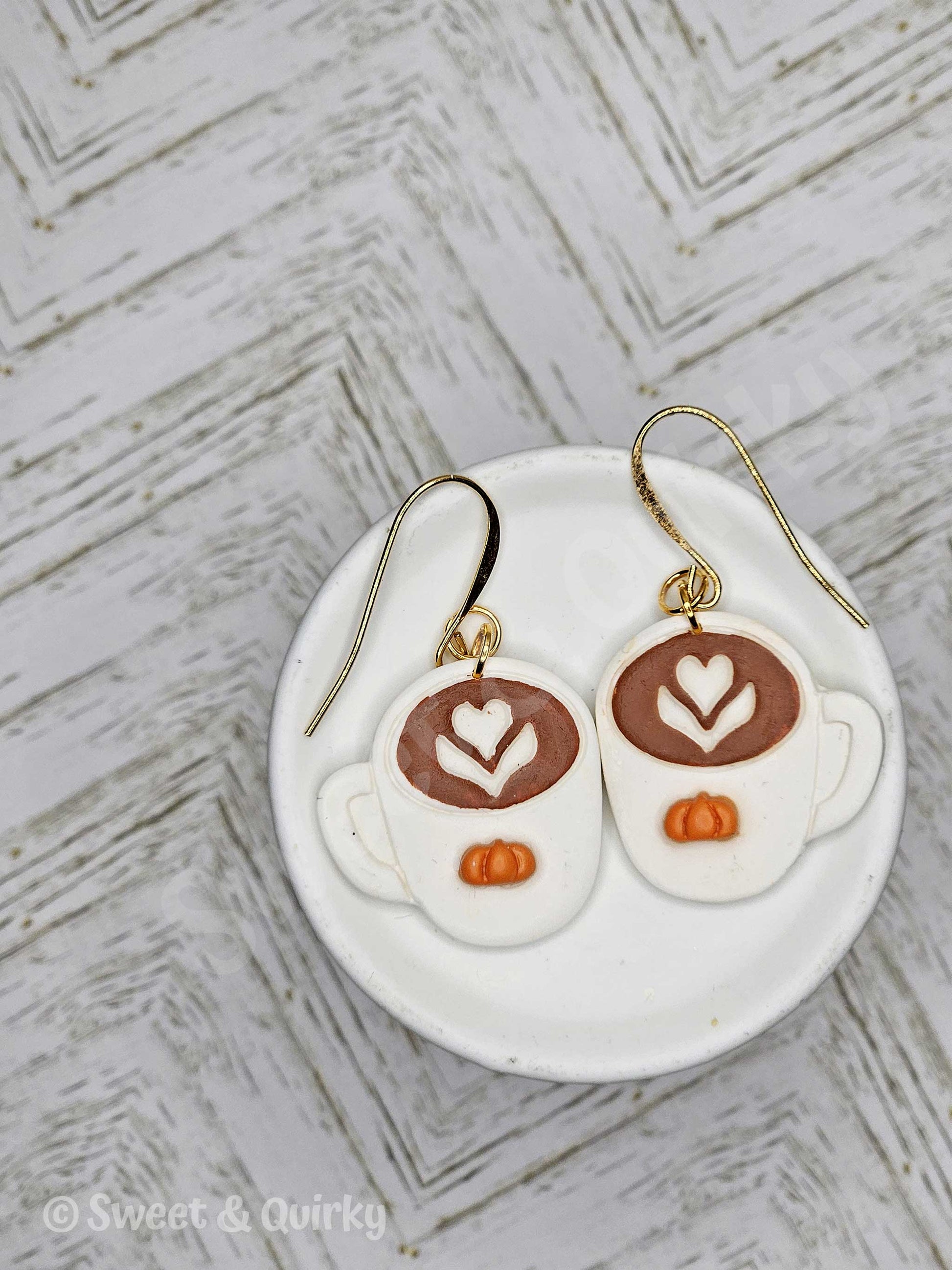 Earrings shaped like coffee cups with latte art design on a white dish with a textured background.
