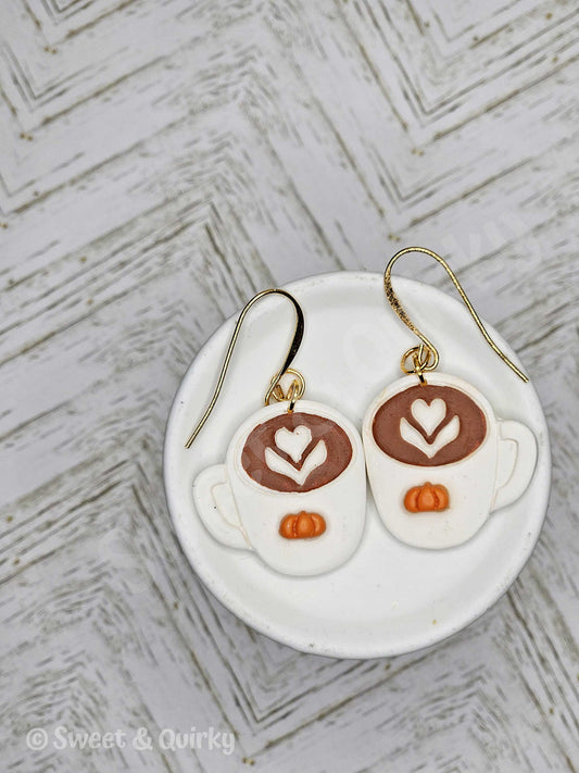 Earrings shaped like coffee cups with latte art design on a white dish with a textured background.