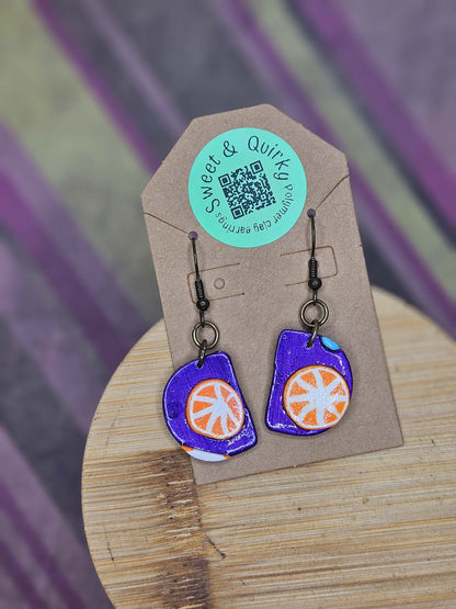 Pair of purple and orange earrings on a Sweet & Quick branded card with a blurred background
