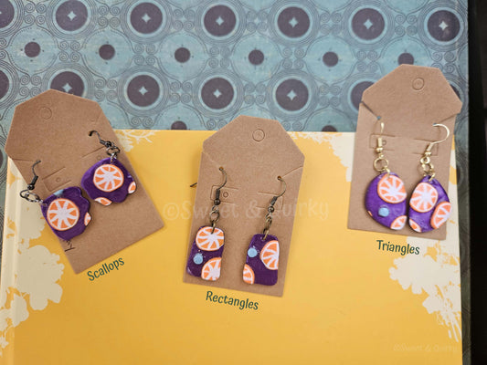 Colorful earrings on brown cards with 'Scallops', 'Rectangles', and 'Triangles' labels against a patterned background.