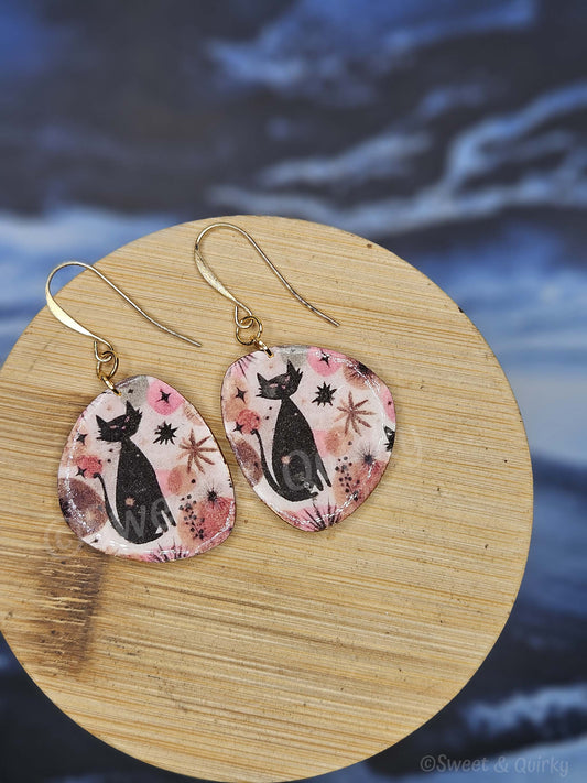 Earrings with black cat design on pink background on wooden board with blue water and cloud background