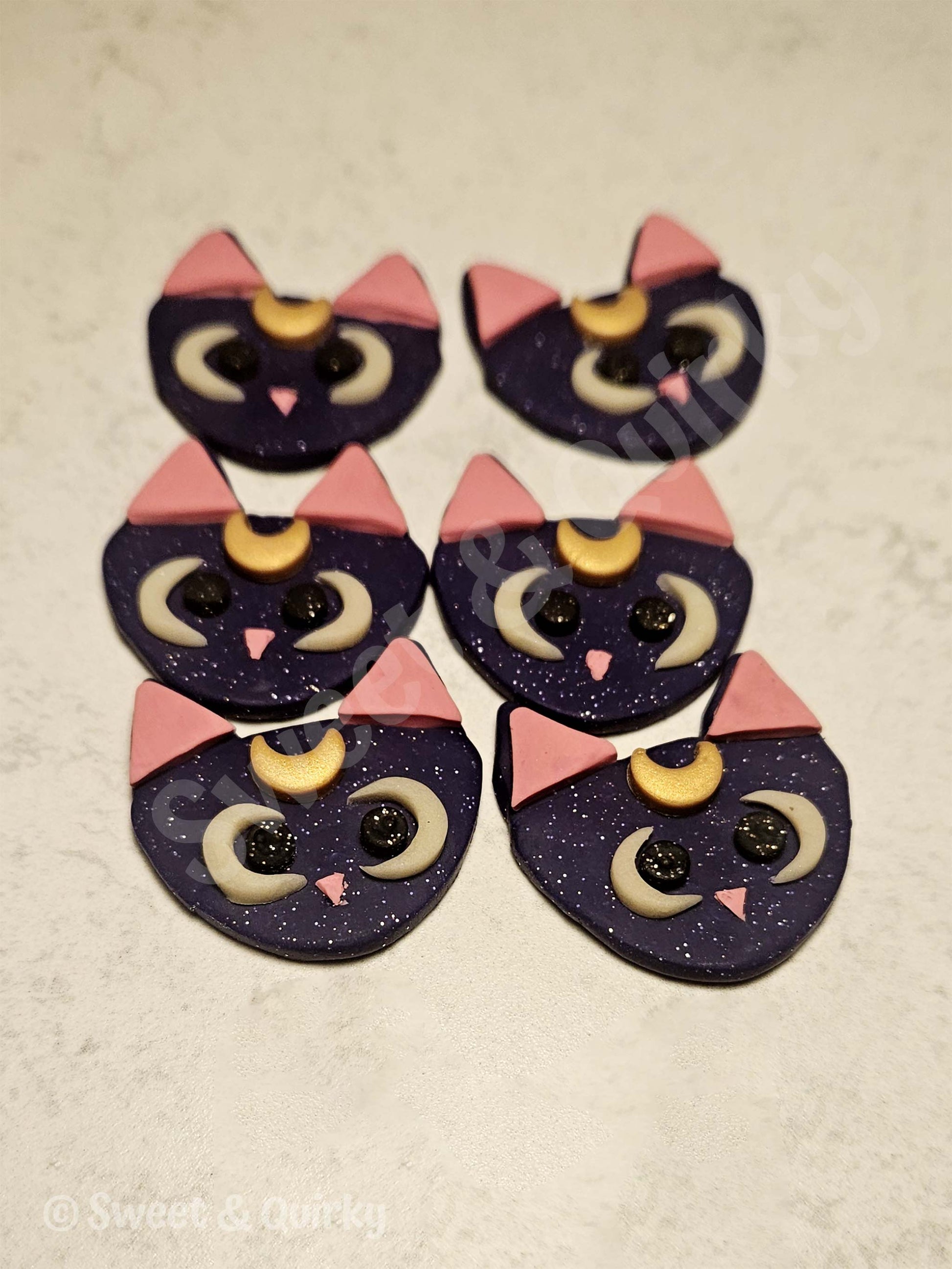 Sailor Moon Luna cat-shaped earrings with purple bodies, pink ears, and black eyes on a beige surface.