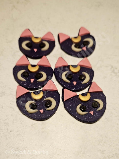 Sailor Moon Luna cat-shaped earrings with purple bodies, pink ears, and black eyes on a beige surface.