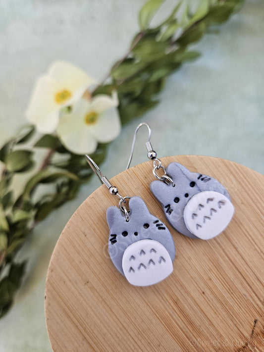 Pair of gray cat-shaped Totoro inspired earrings on a wooden stand with a floral background