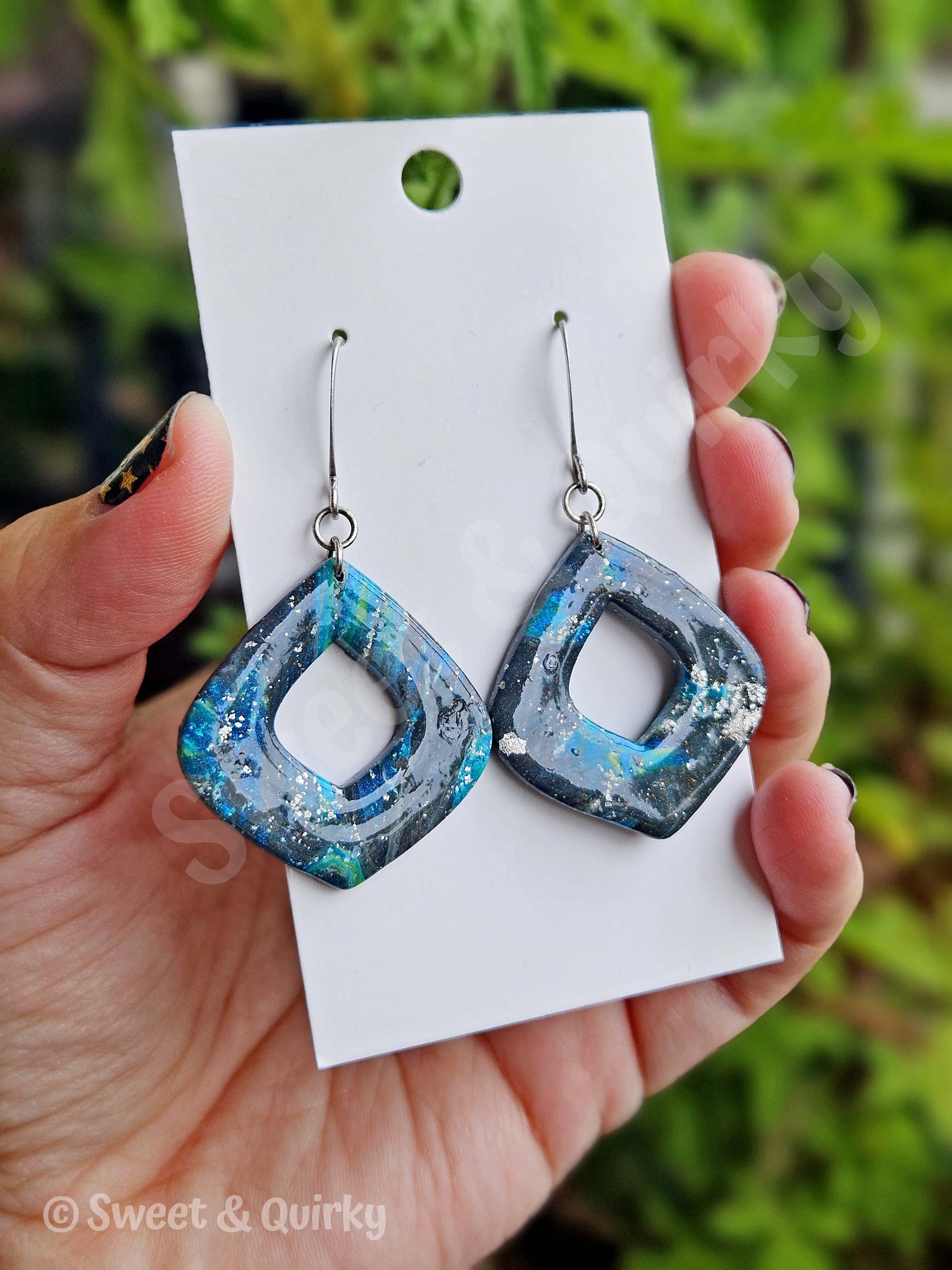Blue and gray earrings on a white card held by a hand with a green leafy background.