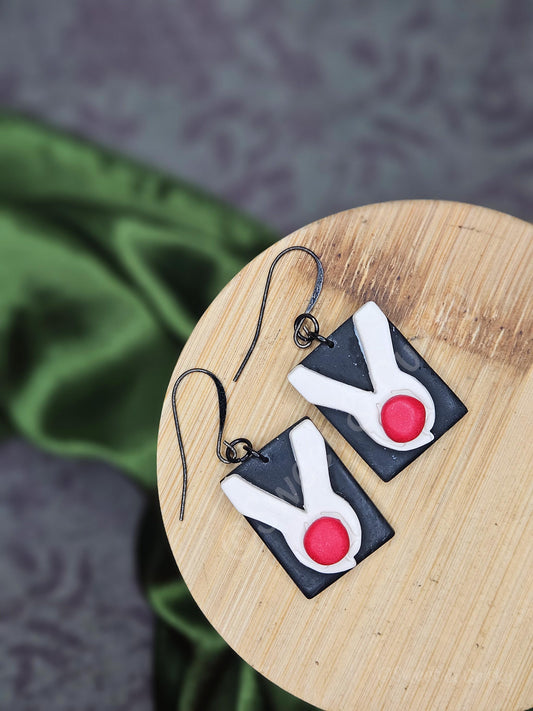 Pair of earrings with black, white, and red design on a wooden surface. Twilight book cover earrings with hands holding an apple.