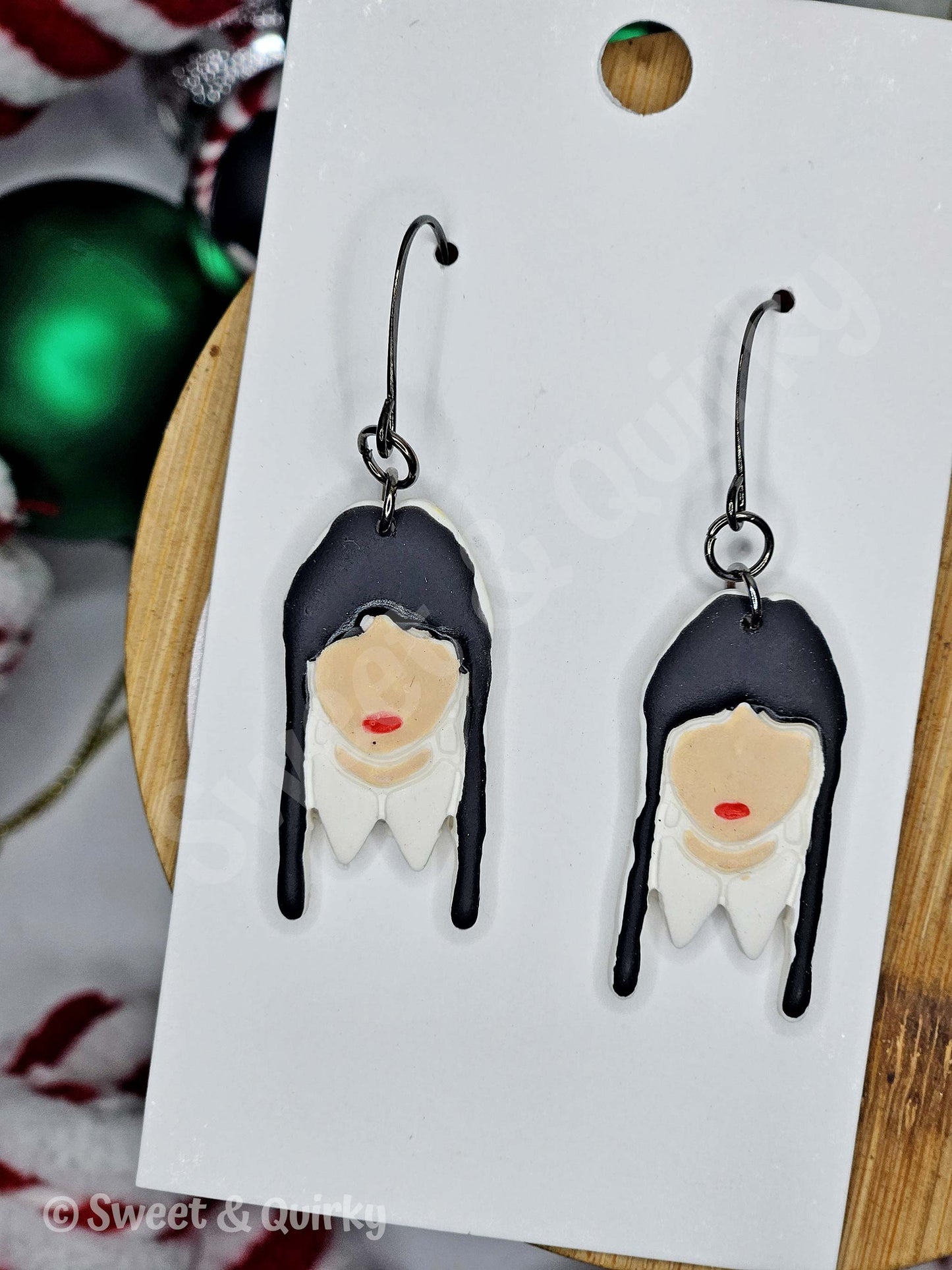 Earrings with Wednesday character design on a white card - black hair with long pigtails and red lips.